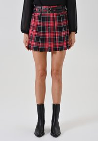 Roter und schwarzer kariierter Minirock mit gefälteltem Design und einem schwarzen Gürtel mit doppelten Metallschnallen. Getragen mit schwarzen Stiefeletten.
