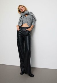 Kvinde i grå knaplukket cropped sweater med matchende tørklæde, sorte brede faux læderbukser og sorte sko med firkantet tå, læner sig op ad hvid væg.