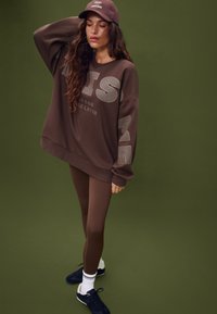 Felpa marrone con grande testo grigio, abbinata a leggings marroni coordinati. Scarpe da ginnastica nere con strisce bianche e un cappellino marrone. Sfondo uniforme.