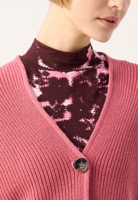 Rožinė megzta kardiganas su V formos iškirpte ir viena saga, uždėtas ant rudos, su rožiniu abstrakčiu raštu.