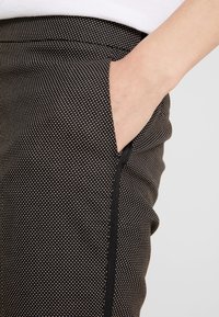 Pantalons noirs en tissu texturé avec de petits pois blancs, une poche latérale et un design épuré avec un biais noir le long du bord.