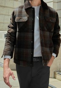 Uomo che indossa un giaccone a quadri marrone scuro con collare in shearling sopra una camicia bianca, con una mano in tasca, in piedi all'aperto accanto a un edificio di pietra.