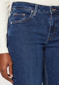 Jeans in denim blu scuro con vestibilità slim, design classico a cinque tasche, cuciture a contrasto e un piccolo logo tricolore sulla tasca anteriore.