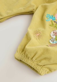 Manche d'un vêtement en velours côtelé jaune avec un poignet élastique et une broderie florale en bleu, rose, vert et des patchs en tissu à motifs.