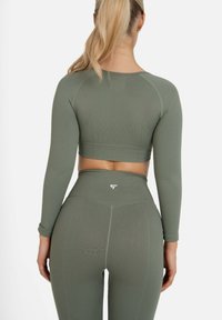 Žena s plavom repom koja nosi usklađeni maslinasto zeleni dugi cropped top s teksturom i helanke visokog struka, stojeći okrenuta leđima.
