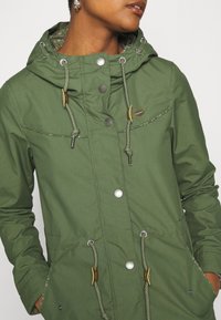 Grön huva jacka med silverfärgade tryckknappar och dragsnören. Har ett strukturerat tyg och ett mönstrat innerfoder. Två framfickor.