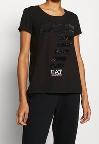 Černé tričko s krátkými rukávy, zdobené flitrovanými pruhy a logem EA7 Emporio Armani na přední straně. Měkký bavlněný materiál.