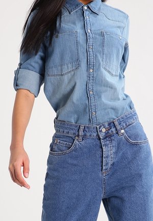 Kobieta ubrana w jasną, dżinsową koszulę zapinaną na guziki z podwiniętymi rękawami oraz wysokiej talii niebieskie dżinsy na jednolitym tle.