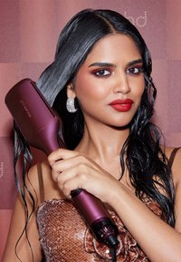 Haarglätter in tiefem Purpur, ausgestattet mit einer breiten Platte zum Stylen. Modell mit nassen Haaren, trägt ein braunes Pailletten-Oberteil und silberne Ohrringe.
