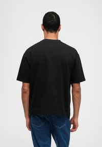 Μαύρο βαμβακερό t-shirt με κοντά μανίκια, στρογγυλή λαιμόκοψη και άνετη εφαρμογή, εικονιζόμενο από πίσω. Συνδυάζεται με μπλε τζιν παντελόνι.