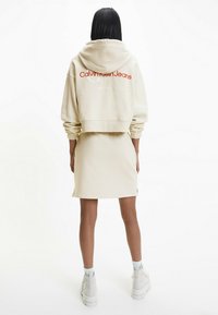 Sudadera beige recortada con texto rojo "Calvin Klein Jeans", combinada con una falda beige a juego. Textura suave, corte holgado y aberturas laterales.