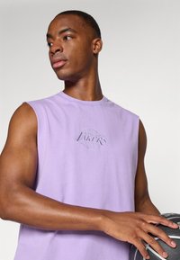 T-shirt sans manches violet arborant le logo "Los Angeles Lakers" en gris brodé. Le mannequin tient un ballon de basket, mettant en valeur des bras musclés.
