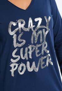 Donkerblauwe longsleeve shirt gemaakt van zachte stof, met zilver metallic tekst: "RAAR IS MIJN SUPERKRACHT," met een V-hals ontwerp.