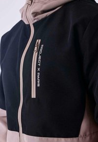 Chaqueta negra y beige con un bolsillo con cremallera. El material parece texturizado y presenta el logo impreso "PROJECT X PARIS" en la parte frontal.