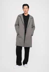 Manteau gris léger avec capuche, boutons et poches latérales, superposé à un pull noir texturé et un pantalon noir ample, associé à des baskets blanches.