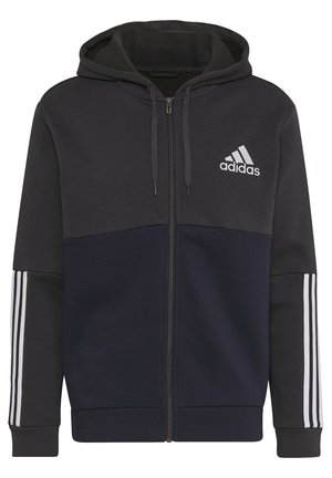 Mężczyzna w czarnej i ciemnoniebieskiej bluzie z kapturem Adidas na zamek błyskawiczny z trzema białymi paskami na rękawach oraz pasujących ciemnoniebieskich spodniach.