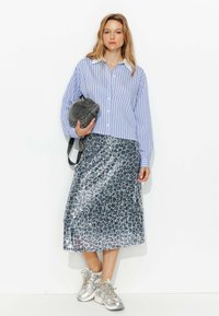 Chemise à rayures bleues et blanches avec boutons, jupe argentée pailletée, sac gris et chaussures de sport argentées. La tenue présente une coupe ample et des textures brillantes.