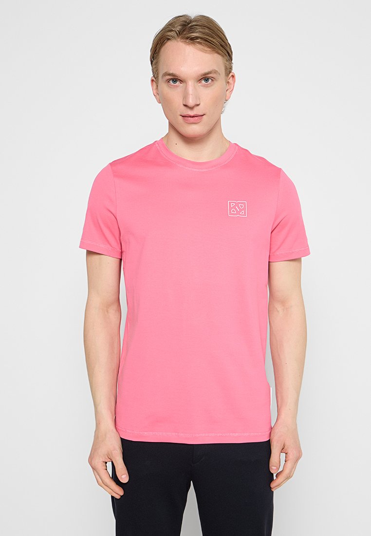 Młody mężczyzna z jasnymi włosami, ubrany w prostą różową koszulkę z krótkim rękawem z małym białym logo na lewej piersi, stojący na tle białego tła.
