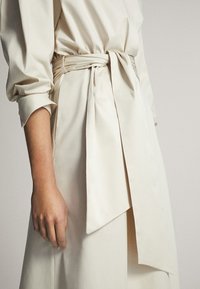 Robe couleur crème avec une ceinture nouée, manches longues et texture douce. Présente un design fluide avec des plis le long du corps.