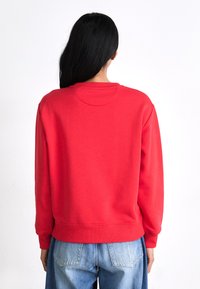 Red crew-neck sweatshirt with a relaxed fit, ribbed cuffs, and hem. Smooth fabric texture; worn with high-waisted light blue denim.