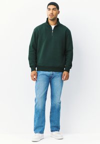 Tmavě zelená mikina na zip s žebrovanými manžetami a lemem, v kombinaci s světle modrými džínami a bílými teniskami. Hladká textura látky.