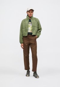 Zelená chlupatá bunda na zip, tričko s grafikou, hnědé kalhoty a šedé boty. Model má také světlou čepici a stojí před bílým pozadím.