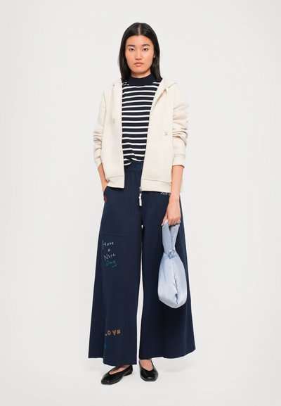 Woman wearing cream hoodie, navy striped shirt, wide-leg navy pants with colorful text, holding light blue handbag, standing against white background.