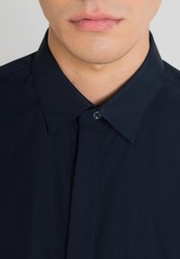 Camicia blu navy con colletto standard, caratterizzata da una trama liscia e una chiusura con un unico bottone sul colletto.