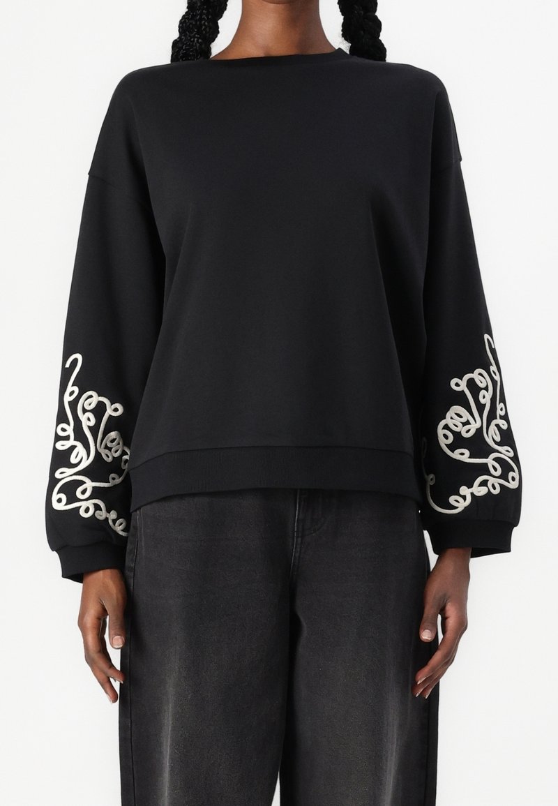 Sweat-shirt noir à manches longues avec broderie abstraite blanche en boucle sur les manches, associé à un pantalon ample gris foncé.
