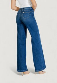 Siniset leveälahkeiset farkut, joissa korkea vyötärö, neljä taskua ja näkyvät ompeleet. Kangas on sileäpintainen ja teksturoitu.