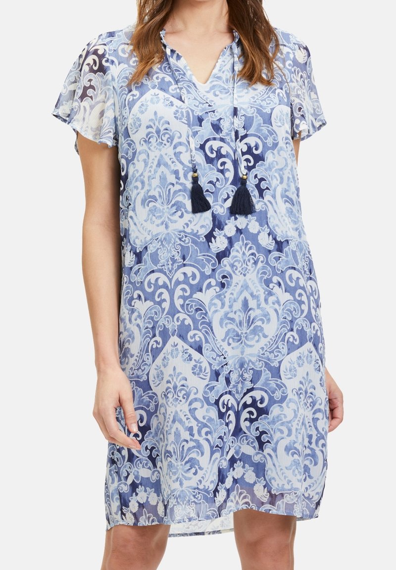 Krátký rukáv, modro-bílý vzorovaný šaty s V-výstřihem a třásňovými akcenty. Lehká, splývavá látka s květinovým damaskovým vzorem.