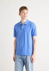 Blaues Baumwoll-Polohemd mit kurzen Ärmeln, einem Kragen und einem kleinen gestickten Logo auf der linken Brust. Getragen mit hellblauen Jeans.