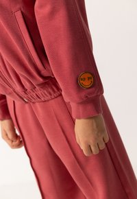 Kind draagt een rode trainingsbroek en een bijpassend sweatshirt met een kleine oranje glimlachend gezichtje patch nabij de pols op de mouw.