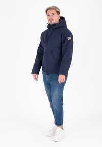 Giacca impermeabile blu navy con cappuccio, dotata di chiusura con zip, tasche laterali e una patch con logo sulla manica sinistra.