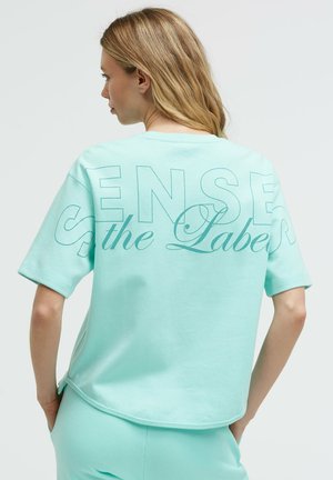 Frau mit langen blonden Haaren, die ein locker sitzendes hellblaues T-Shirt und Hose trägt. Auf dem T-Shirt steht in großen Buchstaben "Sense the Label".