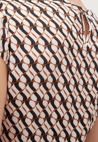 Blouse à motifs en tissu léger présentant un design géométrique en noir, blanc et rouille, avec un détail clé à l'arrière.