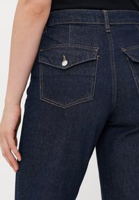 Close-up of person wearing dark blue denim jeans with flap pockets secured by silver buttons and visible orange stitching.