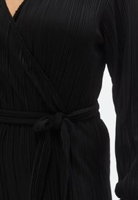 Juoda klostuota suknelė su diržu-rankogaliu. Ji pasižymi giliau įpjauta V formos iškirpte ir ilgomis rankovėmis, pagaminta iš lygios medžiagos su smulkiomis vertikaliomis tekstūromis.