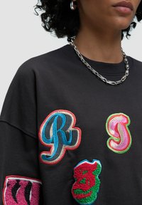 Il felpa nera in cotone presenta colorati ricami a toppe di forme varie: "R" blu, "J" rosa, "3" verde e strisce rosa sulla manica.