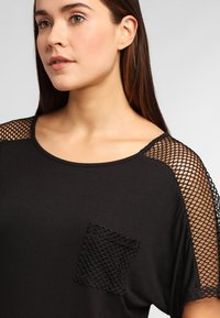 Zwarte top met korte mouwen, voorzien van gaasdetails en een gestructureerde, diamantvormige zak. Gladde stof met een relaxte pasvorm.