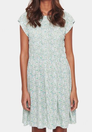 Kvinde iført en kortærmet, knælængde kjole med lys grøn og blå blomsterprint og en detaljeret talje.