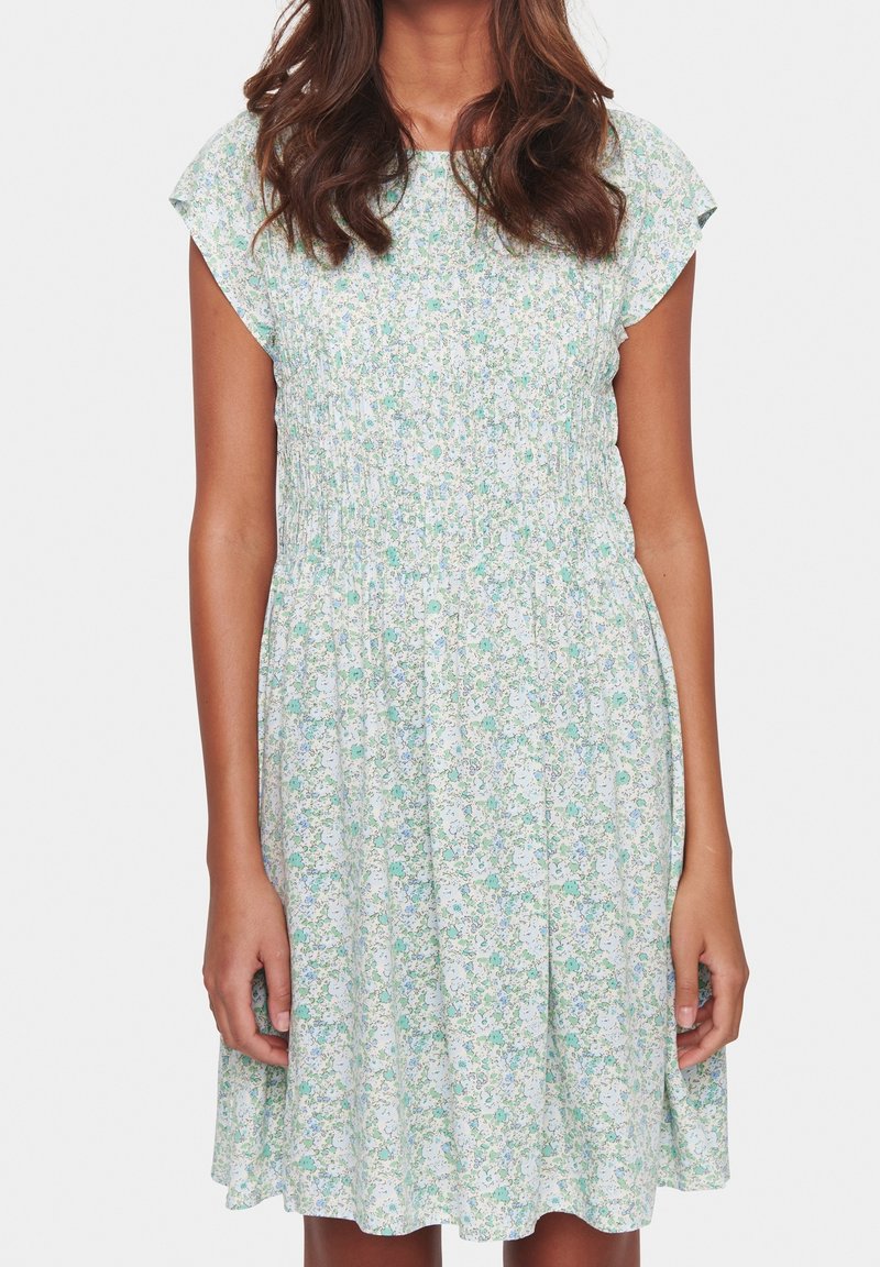 Femme portant une robe à manches courtes, longueur genou, avec un motif floral vert clair et bleu et un détail froncé à la taille.
