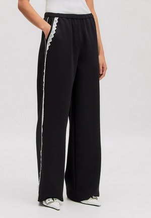 Black wide-leg pants with white lace trim along side seams, worn with white pointed shoes and a white tucked-in top.