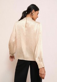 Satin bežs blūze ar garām pūkām, atslēgas caurums aizmugurē un pogājamas piedurknes, saskaņota ar melniem biksēm. Līdzena tekstūra.