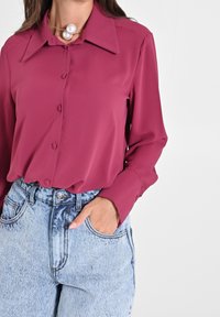 Femme portant une blouse magenta à manches longues boutonnée, rentrée dans un jean taille haute bleu clair délavé, avec une main dans la poche.