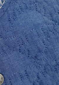 Blauw denimstof met geweven, geborduurde patronen van abstracte vormen. Inclusief een zilveren knop met een sterdetail.