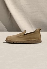 Ταμπά παπούτσι τύπου slip-on με στρογγυλεμένη μύτη, υφασμένο πέλμα, θηλιά έλξης στη φτέρνα, εκτεθειμένο πάνω σε ουδέτερη επιφάνεια με υφασμάτινο φόντο.