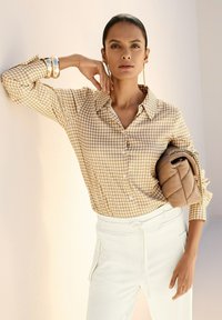 Frau trägt ein beige-weißes Hahnentrittmusterhemd, weiße Hose, hält eine beige gesteppte Handtasche mit goldener Kette, accessorized mit goldenen Armreifen und Ohrringen.