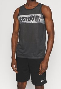 Uomo atletico che indossa una maglietta senza maniche nera con striscia a camo grigio con la scritta "JUST DO IT" e pantaloni corti neri con logo Nike bianco.