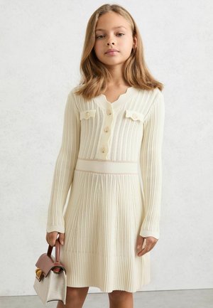 Šaty z rebrovaného úpletu v krémovej farbe s dlhými rukávmi, výstrihom do V, detailmi na gombíkoch a hladkým pásom. Modelka drží malú štruktúrovanú kabelku.